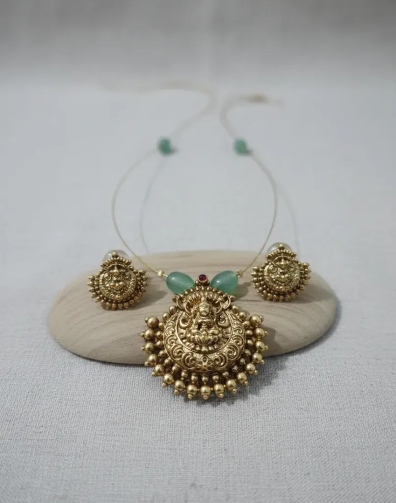 Glamourous gold-finished short haram suite showcasing an elaborate Lakshmi Charm adorned with mint-green gemstones and paired identical filigree earrings.