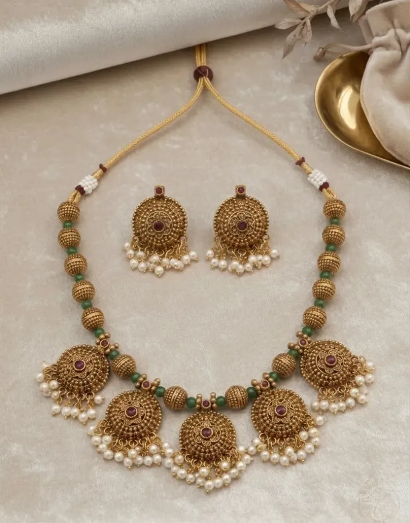 Exude timeless elegance with this antique gold-toned necklace set, featuring intricate circular pendants adorned with ruby-red stones and delicate pearl droplets.