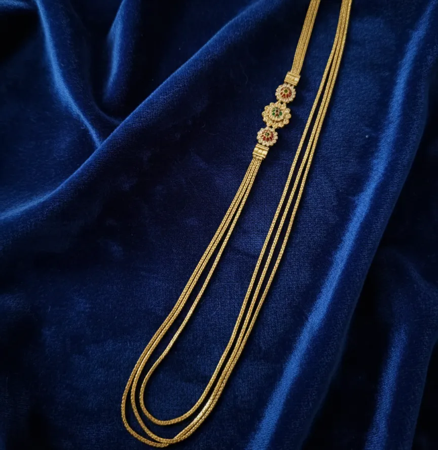 This elegant triple-layered gold long haram features a meticulously crafted floral side-pendant accented with traditional ruby and emerald stones.