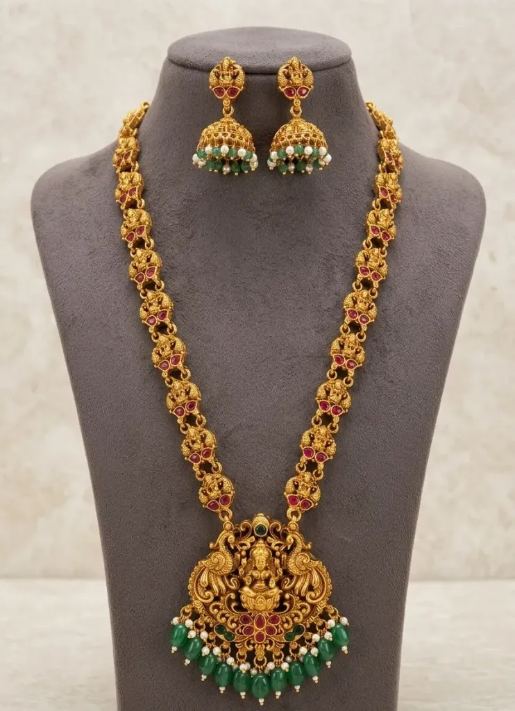 Stunning Gold Toned Jewellery set featuring a Lakshmi Pendent with Red & Green stones, intricate filigree chain and matching jhumka earrings with pearl accents.