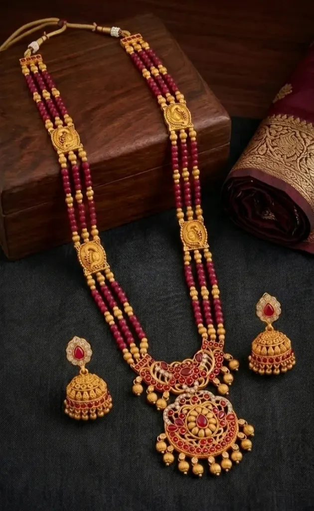 This elegant gold-toned long haram set features three layers of vibrant ruby-red beads accented with intricate peacock-motif side pendants and a majestic centerpiece.